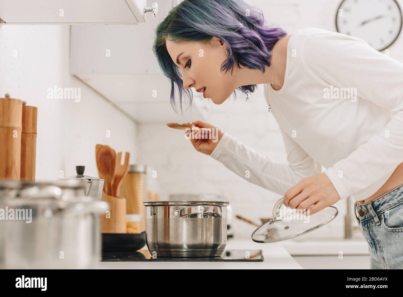 Selective focus of girl with colorful hair preparing food and holding pan lid with spatula near kitchen stove Stock Photo