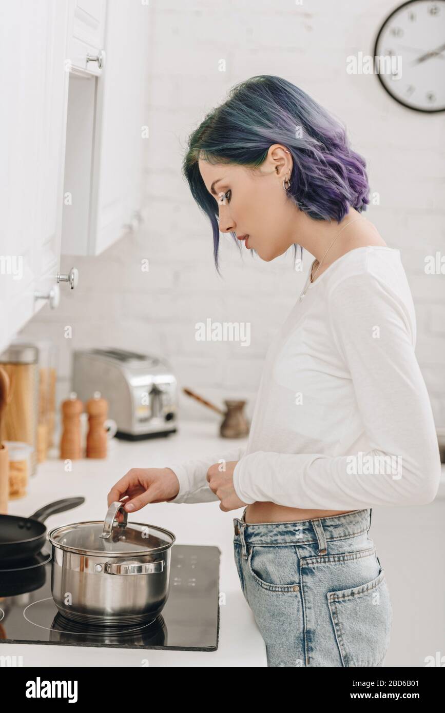 Girl with colorful hair preparing food and touching pan lid near kitchen stove Stock Photo