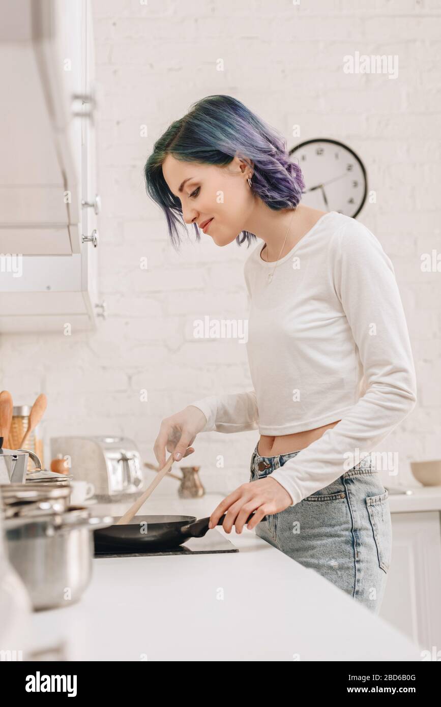 Selective focus of girl with colorful hair preparing food with spatula on frying pan in kitchen Stock Photo