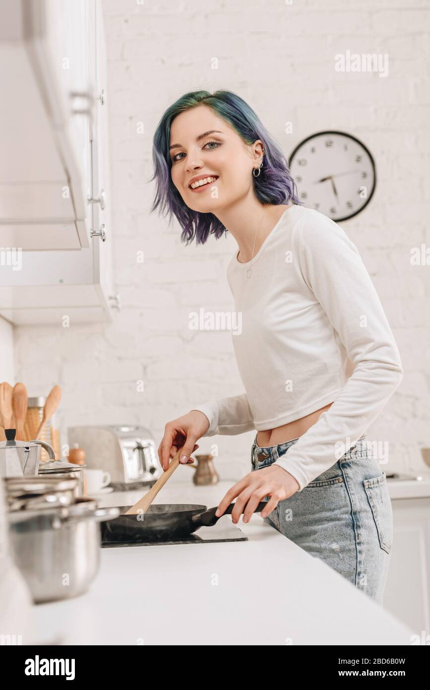 Low angle view of girl with colorful hair preparing food with spatula on frying pan, smiling and looking at camera in kitchen Stock Photo