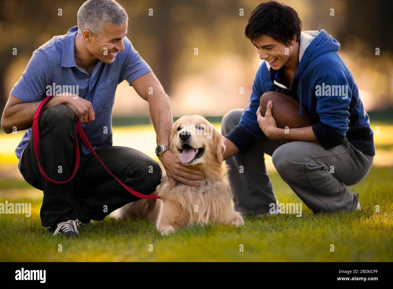 Mature man and his son having fun petting their dog together while crouching in a park. Stock Photo