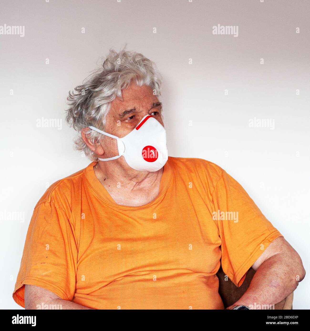 A senior with an antiviral protective FFP3 N99 mask, protecting against coronavirus. White haired Caucasian man in an orange T-shirt on white wall Stock Photo