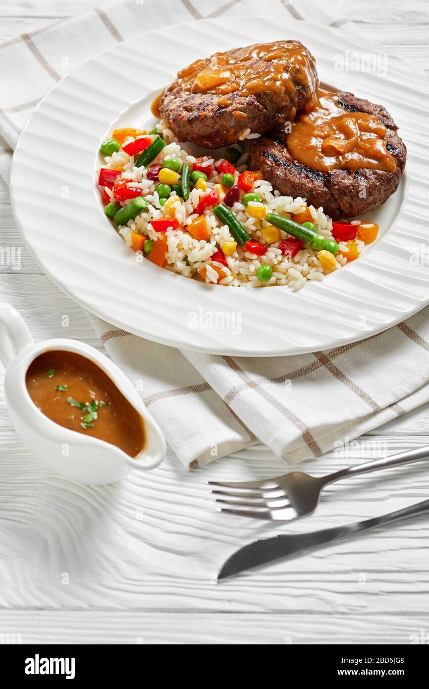 grilled ground beef steaks with onion gravy and rice mixed with vegetables on a white plate on a wooden table, vertical view from above Stock Photo