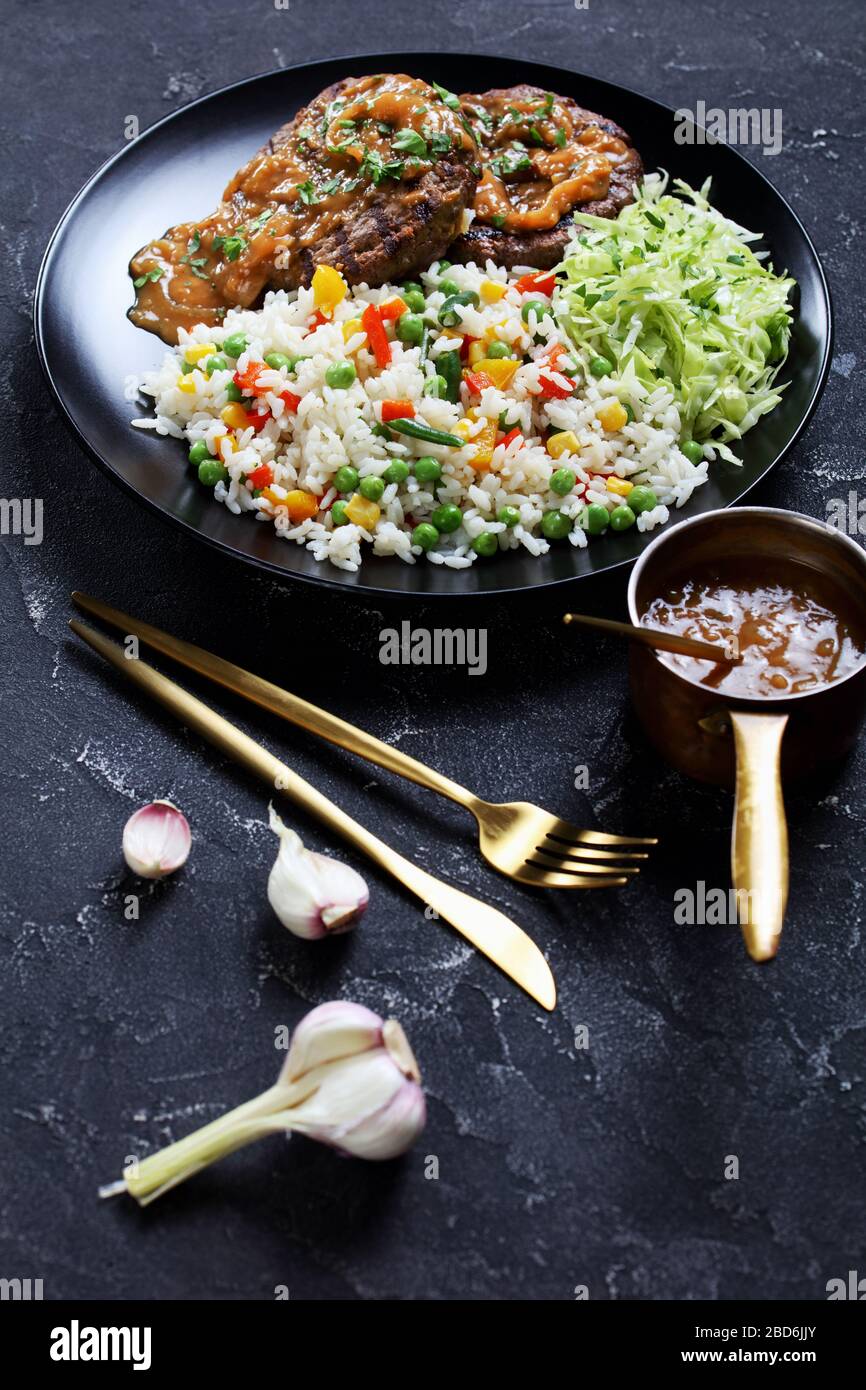 grilled ground beef steaks with onion gravy, coleslaw and rice mixed with vegetables on a black plate on a concrete table, vertical view from above Stock Photo