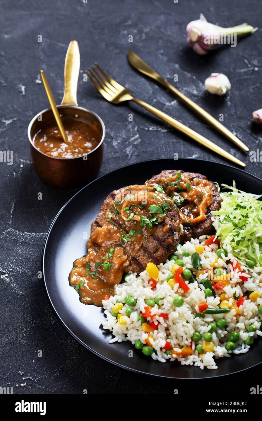 close-up of a portion of grilled ground beef steaks with onion gravy, coleslaw and rice mixed with vegetables on a black plate on a concrete table, ve Stock Photo