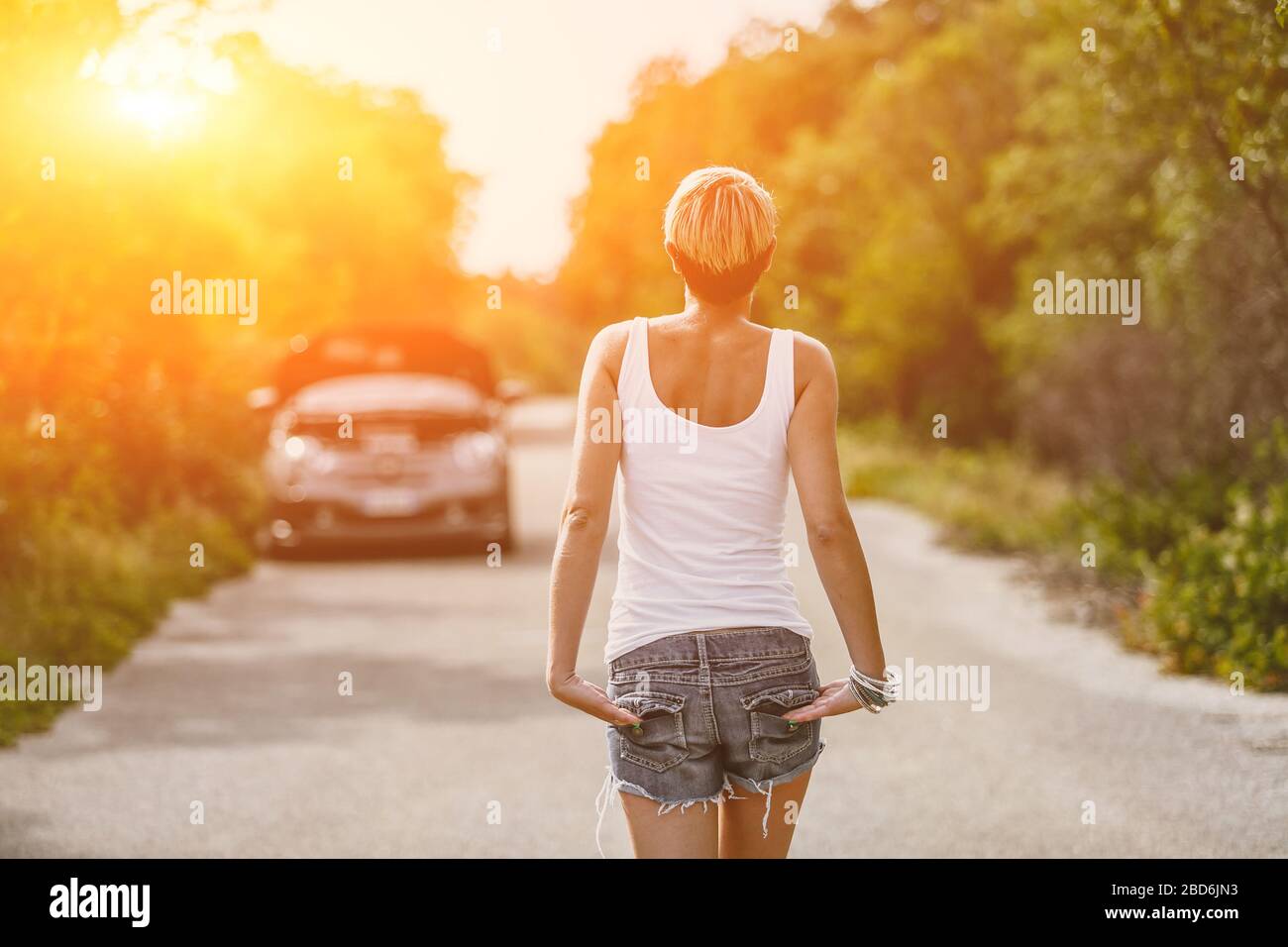 Young sad woman standing near the road by the broken car in the middle of nowhere. Smoke coming out from overheat engine. Waiting car tow service. Stock Photo