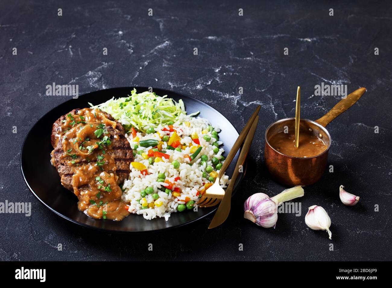 grilled ground beef steaks with onion gravy, coleslaw and rice mixed with vegetables on a black plate on a concrete table, horizontal view from above Stock Photo
