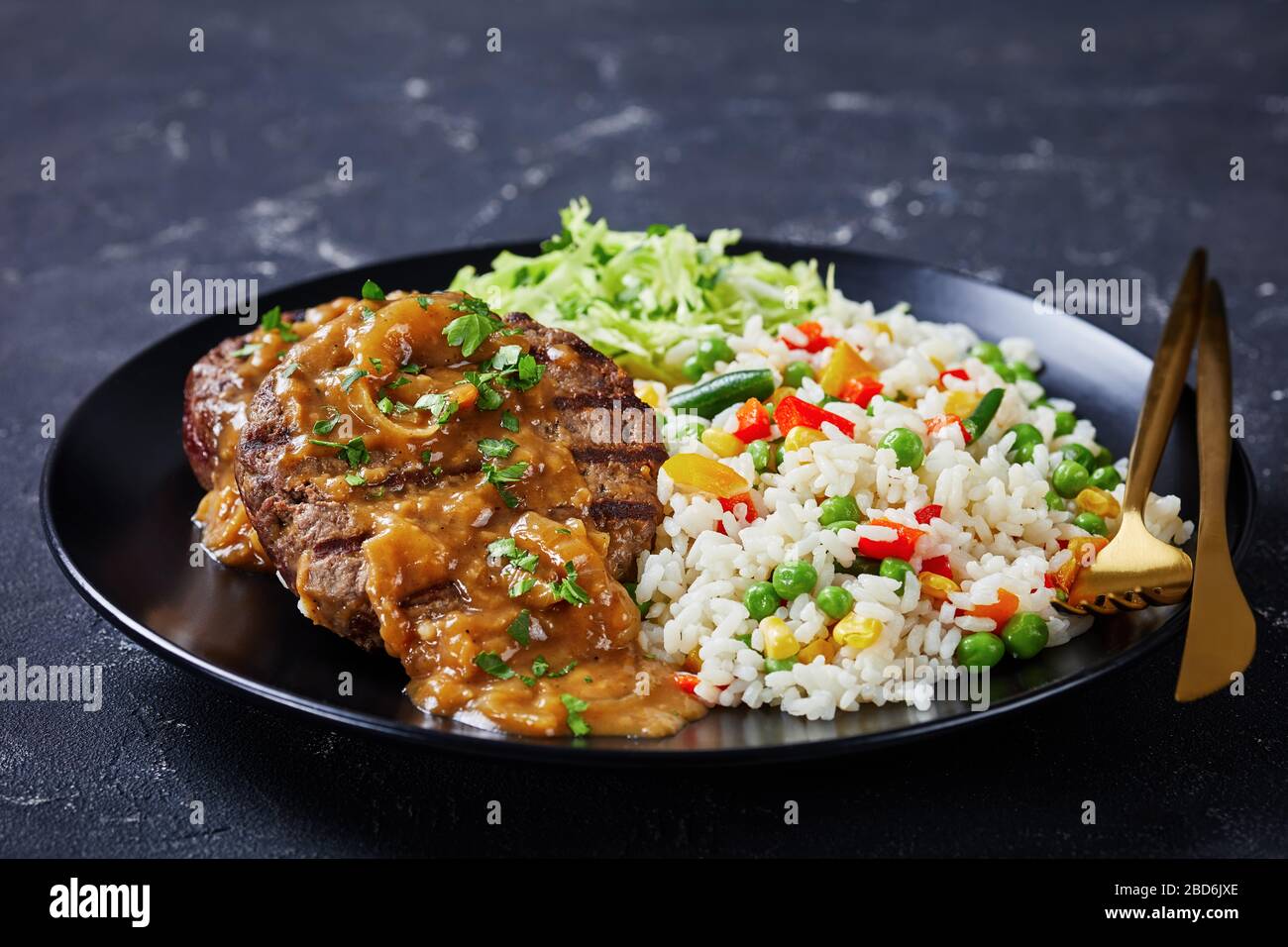 close-up of a portion of grilled ground beef steaks with onion gravy, coleslaw and rice mixed with vegetables on a black plate on a concrete table, ho Stock Photo