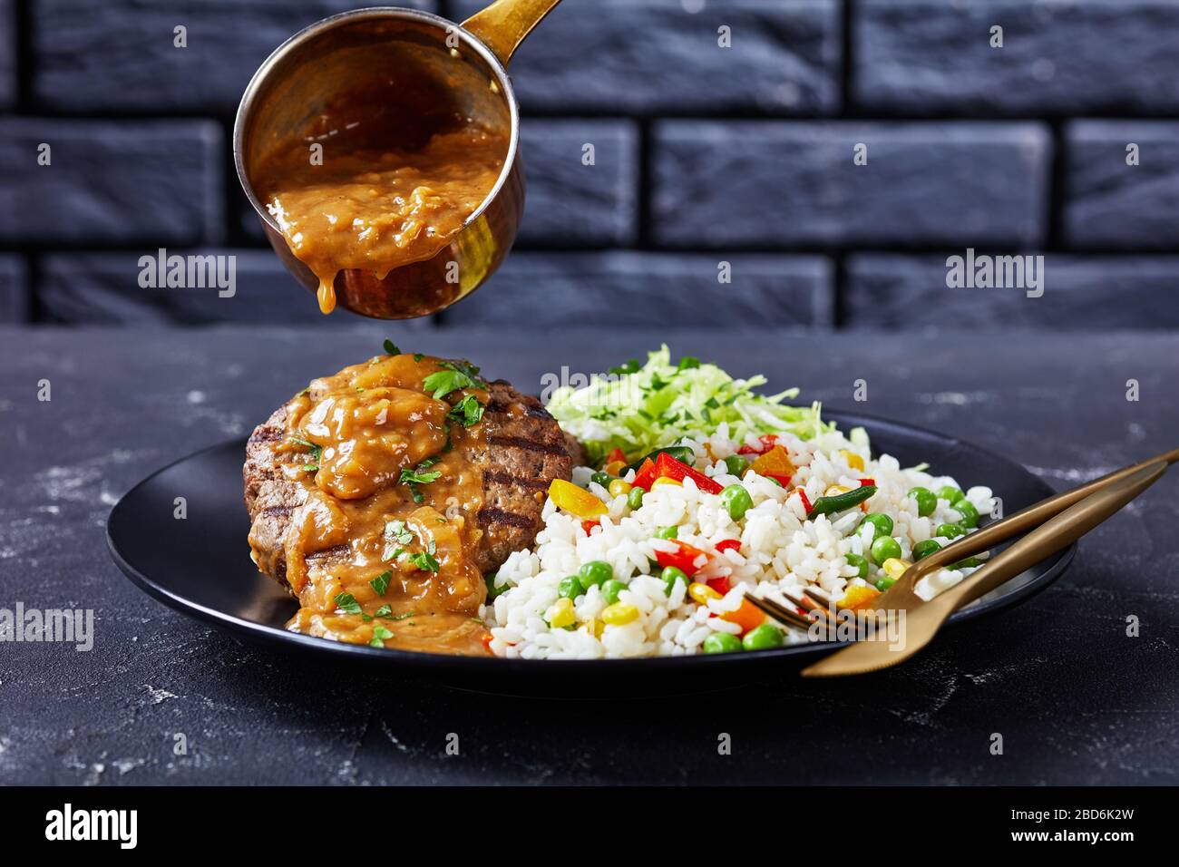 close-up of a portion of grilled ground beef steaks has been pouring with onion gravy, coleslaw and rice mixed with vegetables on a black plate on a c Stock Photo