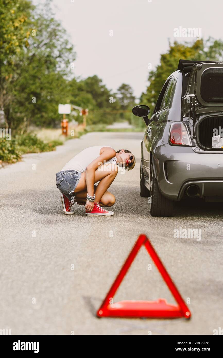 Young beautiful woman spection a rear tire on the side of the road. Car with problems and red triangle to warn other road users. Stock Photo