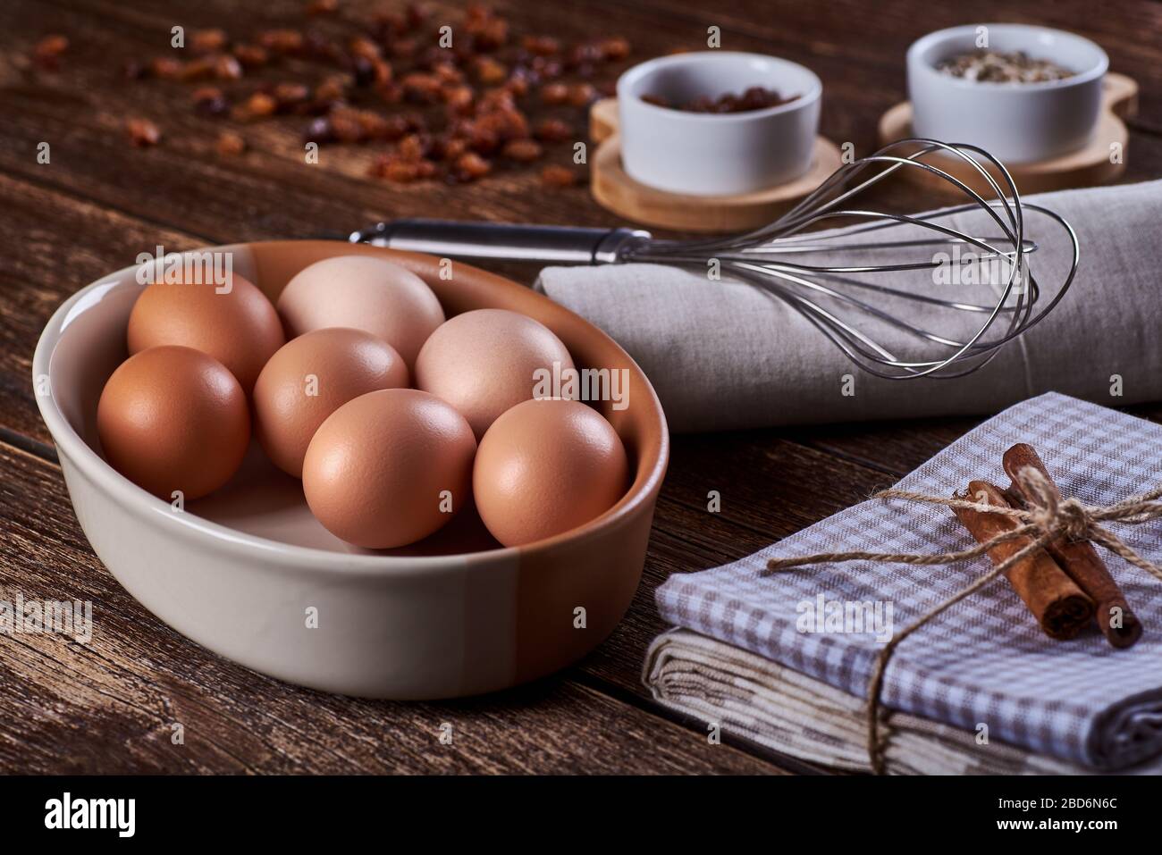 Close-up view of a bowl of hen's eggs, dish-cloth and whisk. Raisins on old wooden board table with space for text. Stock Photo