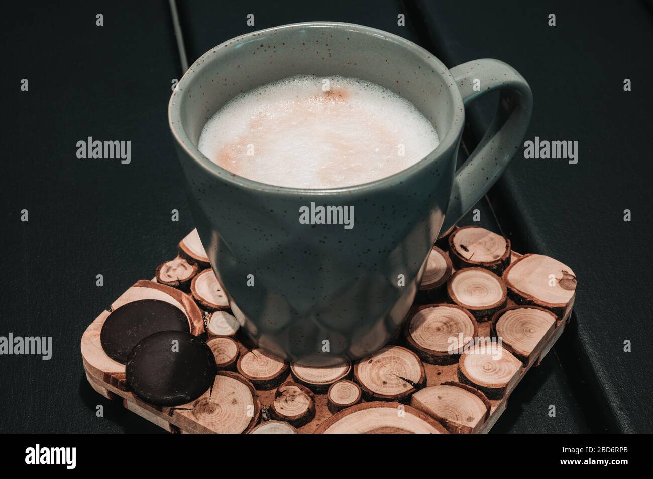 Dark green ceramic cup of latte coffee with milk foam on the wooden textured stand and chocolate candies. Serving beverage in the restaurant or cafe. Stock Photo