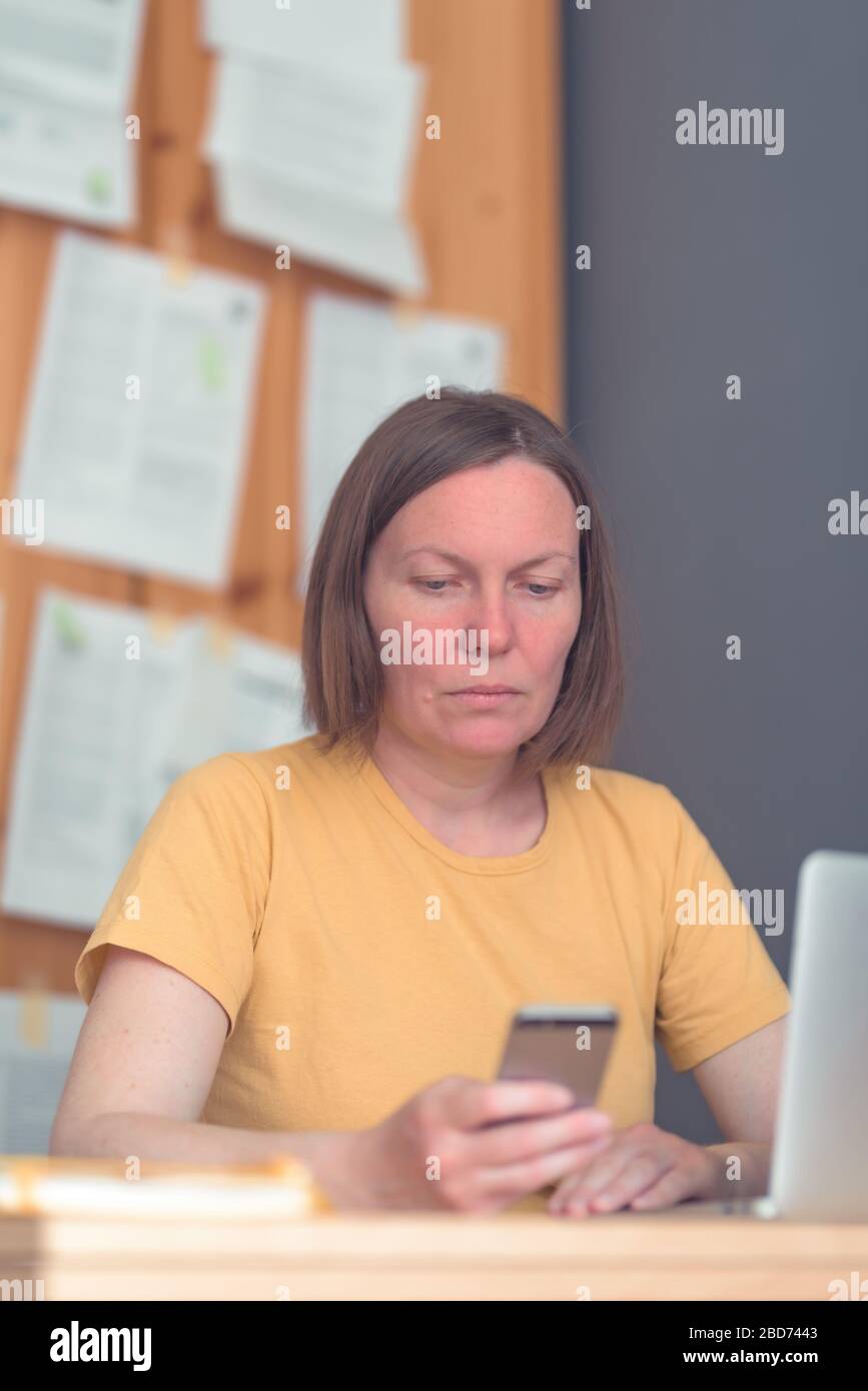 Thoughtful female freelance translator working in home office with laptop computer and smartphone, selective focus Stock Photo