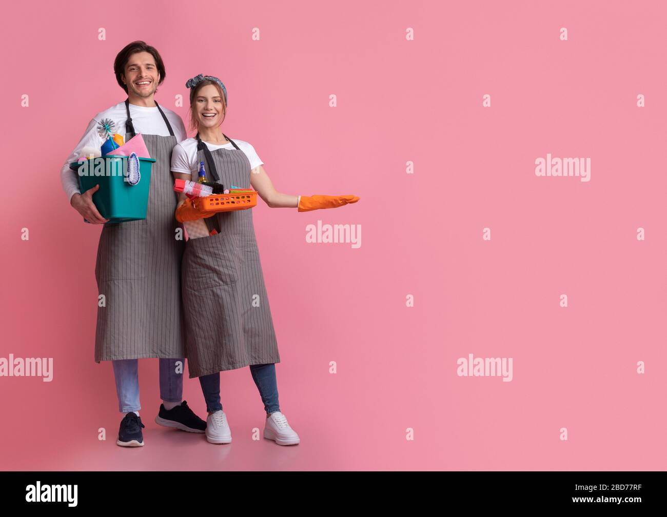 Cleaning Service Team. Smiling Couple Holding Household Supplies And Pointing Aside Stock Photo