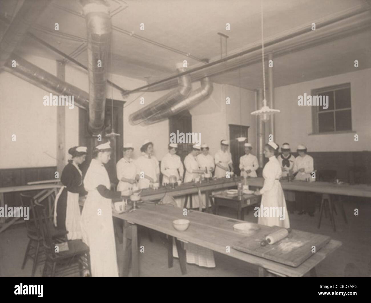 "English: Women receiving cooking instructions in basement of Goderich Library; women standing at U-shaped table; all women wearing white except one who is wearing a black dress with white apron; ventilation pipes come from ceiling into back wall; writing on back in pencil: Goderich Library. Mrs. Felkirk, instructor. Donated by Mrs. Kitty Smith, daughter of Mrs. Murray woman in forefront. Image number 0811-rrs-ogoh-ph; between 1905 (library opened) and 1937 (death of photographer); http://www.historicplaces.ca/en/rep-reg/image-image.aspx?id=14182#i2, larger version here: http://www.sallowsgall Stock Photo