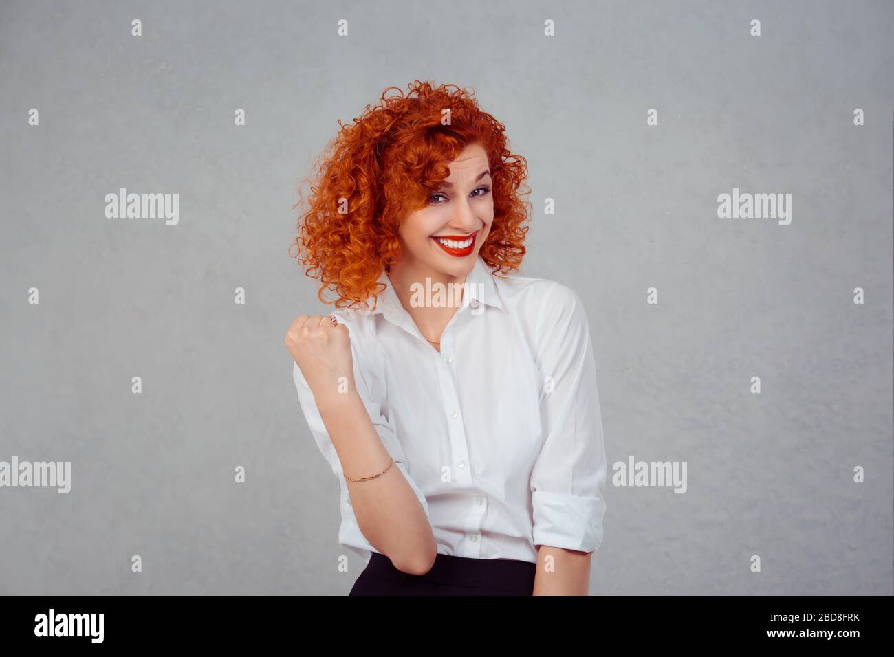 Happy winner. Closeup half body portrait redhead curly woman in white formal shirt exults pumping fists ecstatic celebrates success isolated gray back Stock Photo