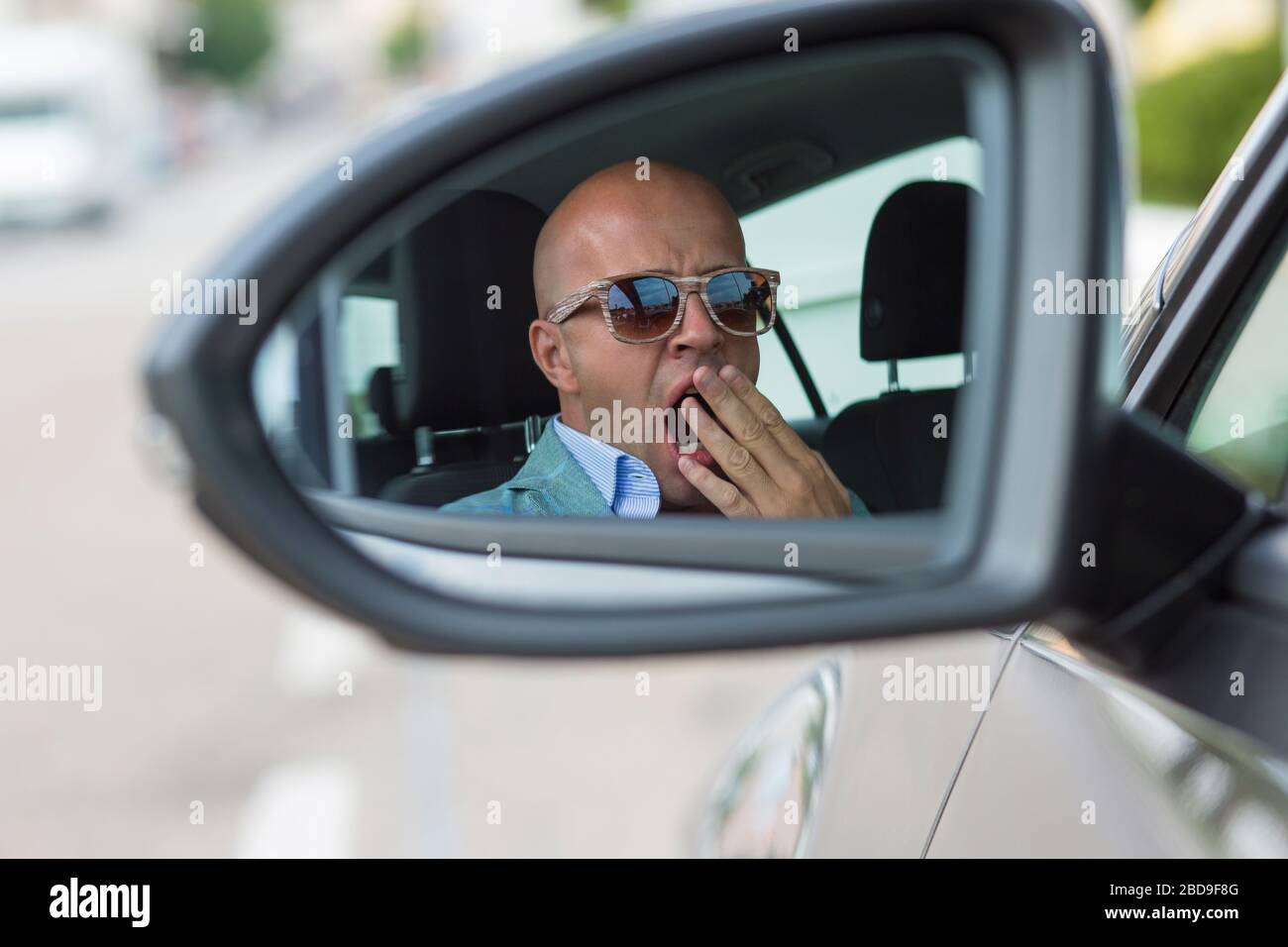 Side mirror view reflection sleepy tired fatigued yawning exhausted young man driving his car in traffic after long hour drive. Transportation sleep d Stock Photo