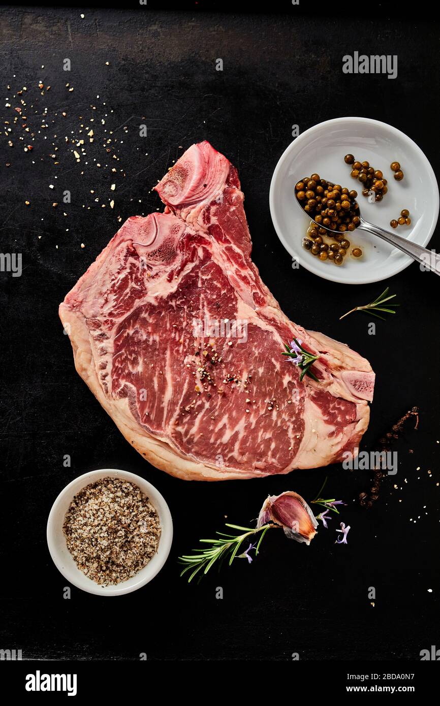 Bone-in raw marbled pork steak with spice rub, fresh herbs and garlic for seasoning in a flat lay on a dark background Stock Photo