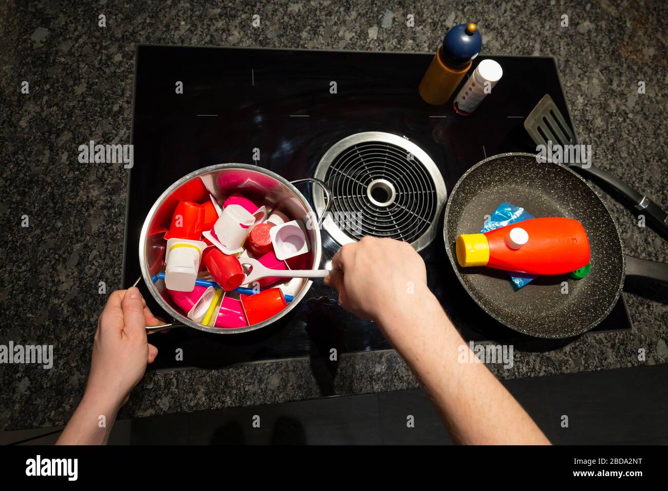 Concept of food pollution with plastic waste. Top view of a persons hands at a stove with a frying and a cooking pan filled with plastic objects Stock Photo