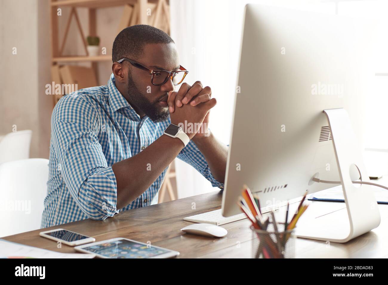 Thinking about business. Thoughtful young afro american man in glasses looking at computer screen while sitting at his working place at home. Working remotely. Freelance. Home office workplace Stock Photo