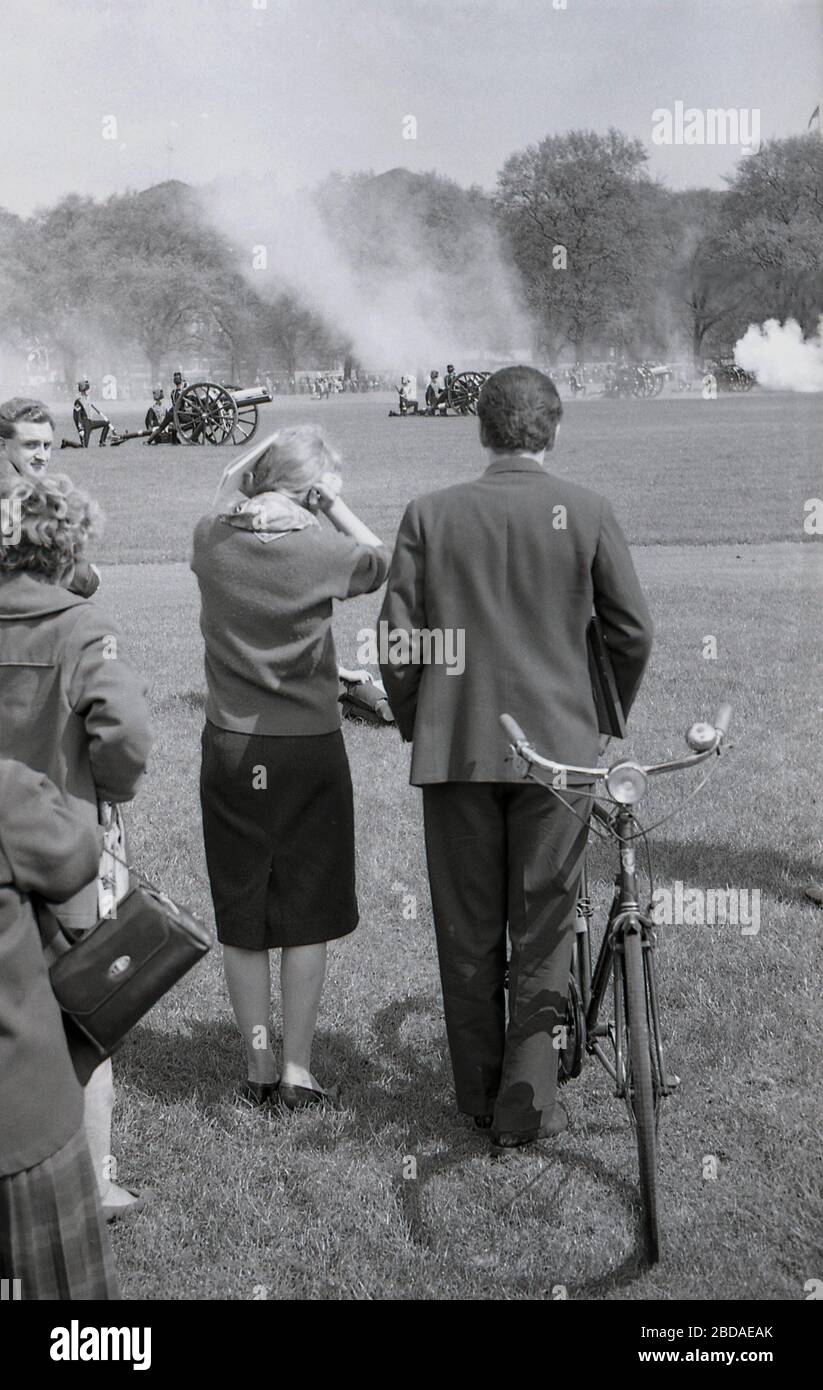 1960s, historical, lady spectator puts her hands on her ears as the sound of the firing of ancient guns at a Royal gun Salute at Green Park near Buckingham Palace, Westminster, London, England. Gun salutes mark special royal occasions including the Queen's birthday and are a tradition display of British pageantry. Stock Photo