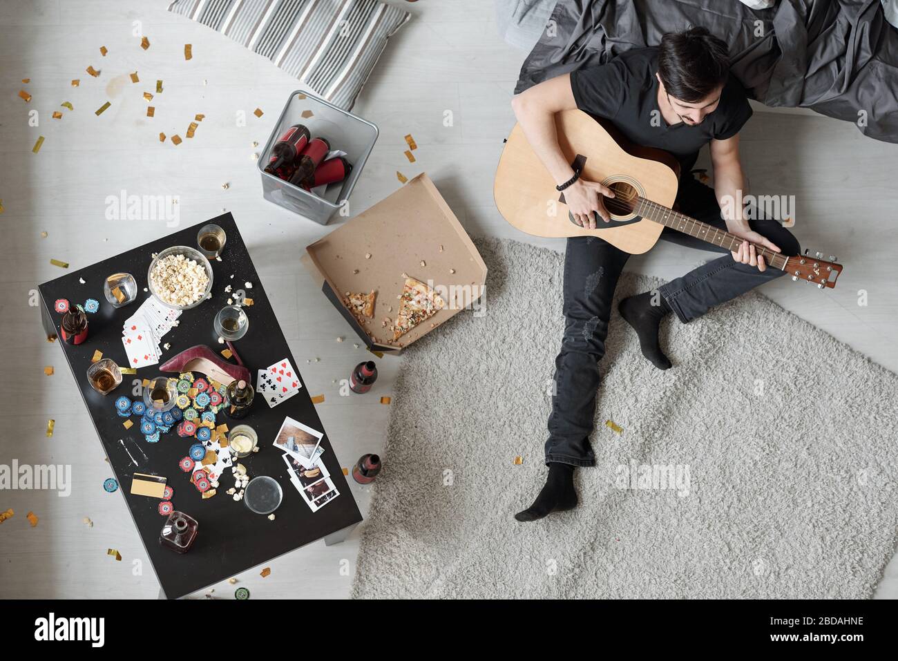 Above view of young man sitting on floor in messy flat after rough poker party and playing guitar while singing song Stock Photo