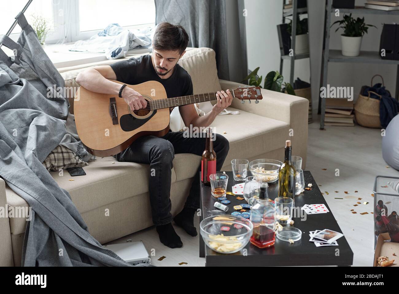Young man sitting on sofa with curtain and playing guitar in messy flat after party, table with alcohol and playing pieces Stock Photo
