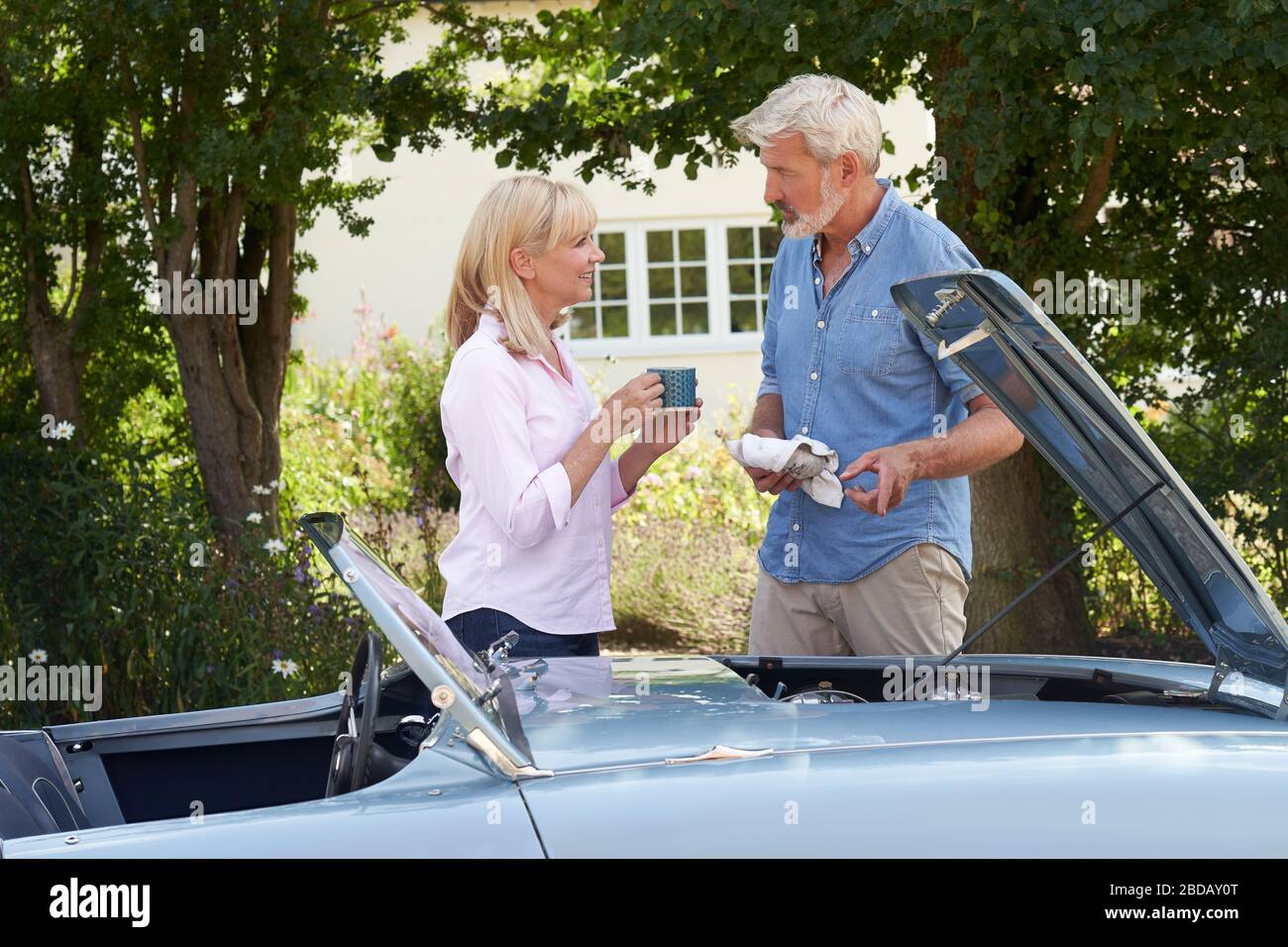 Mature Woman Bringing Hot Drink To Man Restoring Classic Sports Car Working On Engine Under Hood Stock Photo