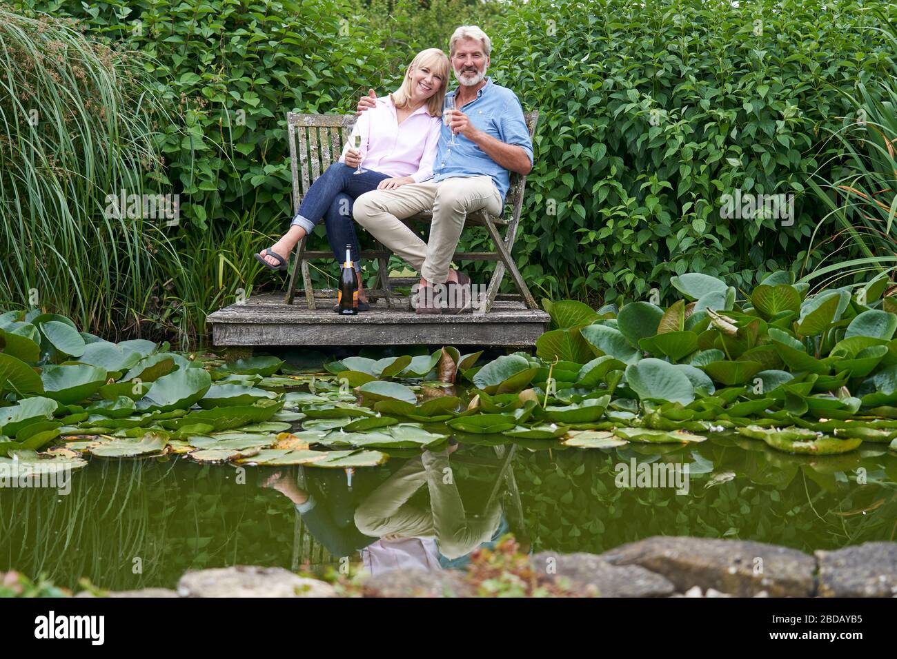 Portrait Of Mature Couple Celebrating With Champagne Sitting On Chairs On Wooden Jetty By Lake Stock Photo