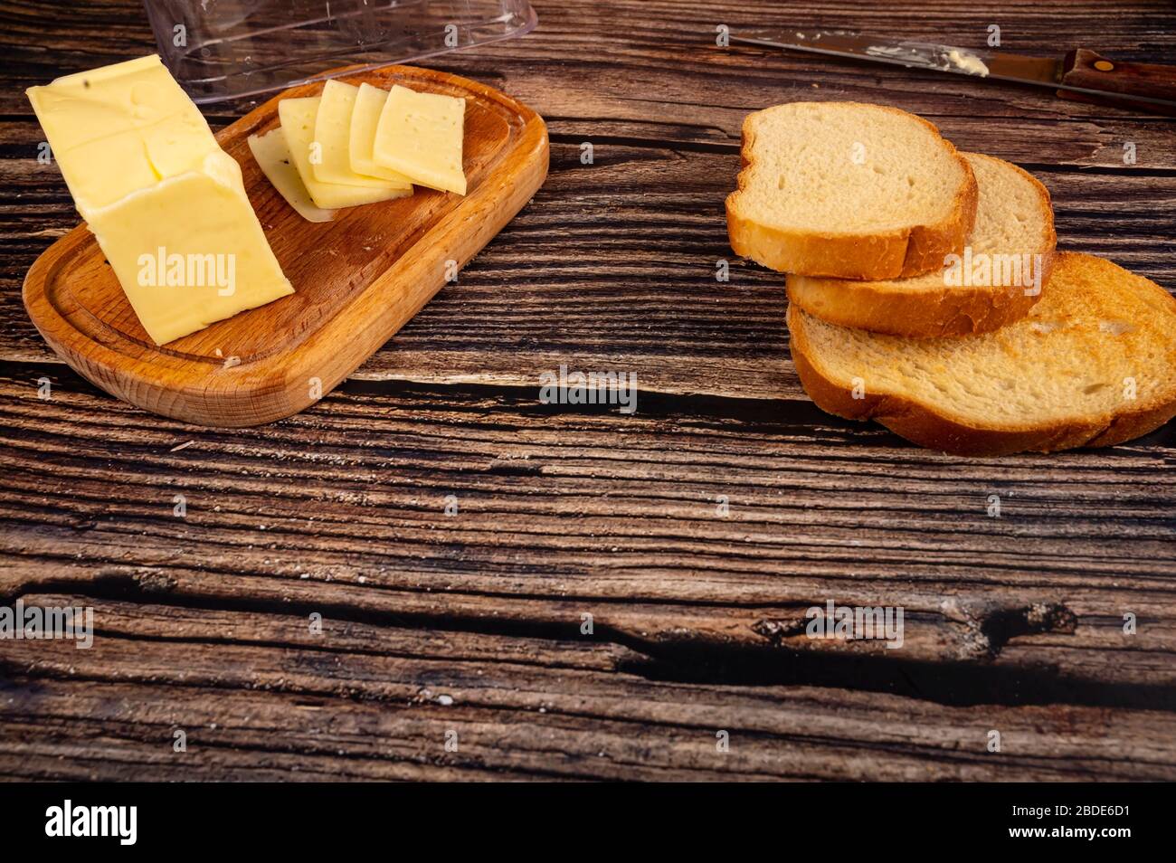 Fresh wheat bread toast and a wooden butter dish with a piece of butter and slices of cheese on a wooden background. Close up Stock Photo