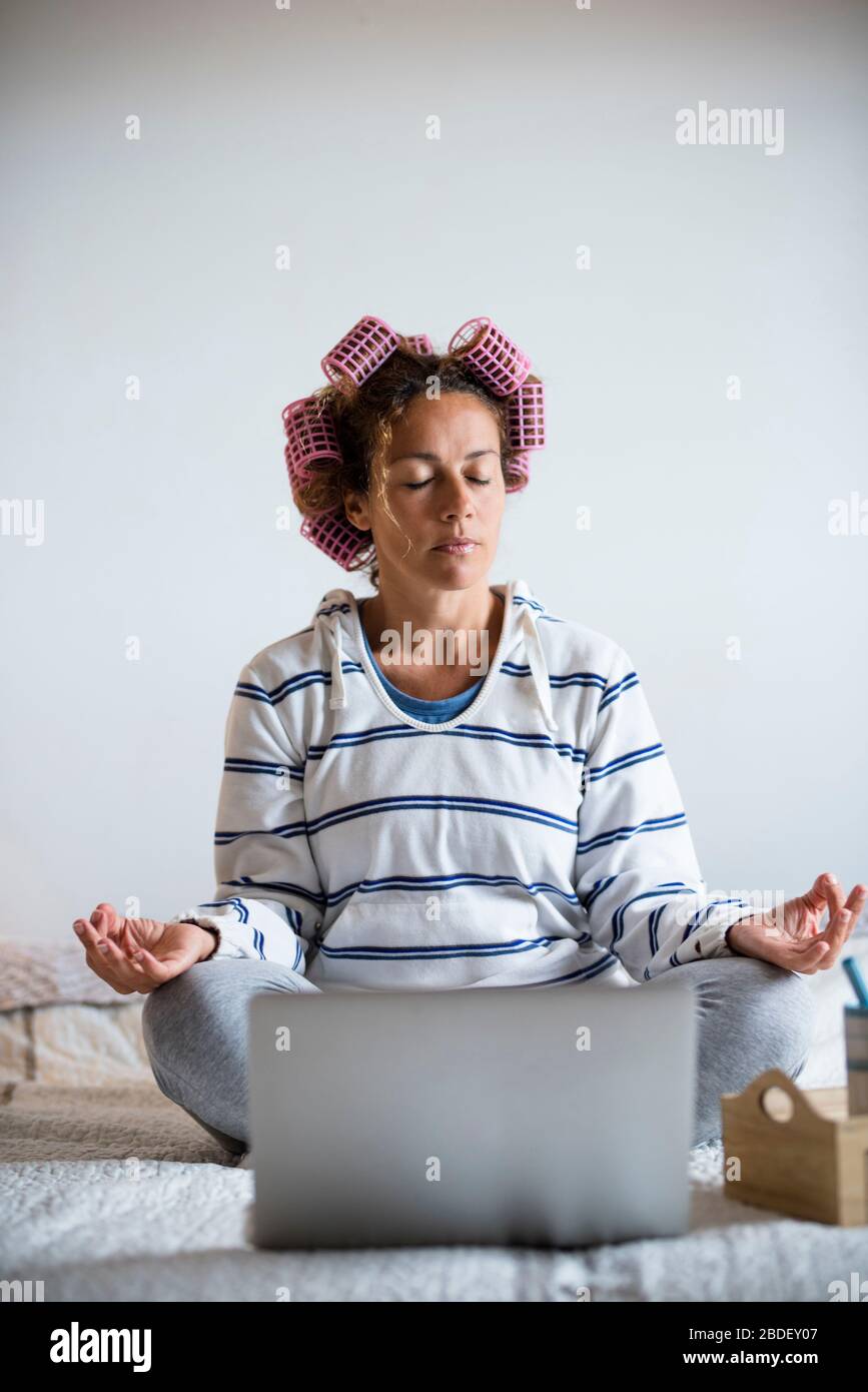 Woman with hair curlers meditating on bed in front of laptop Stock Photo