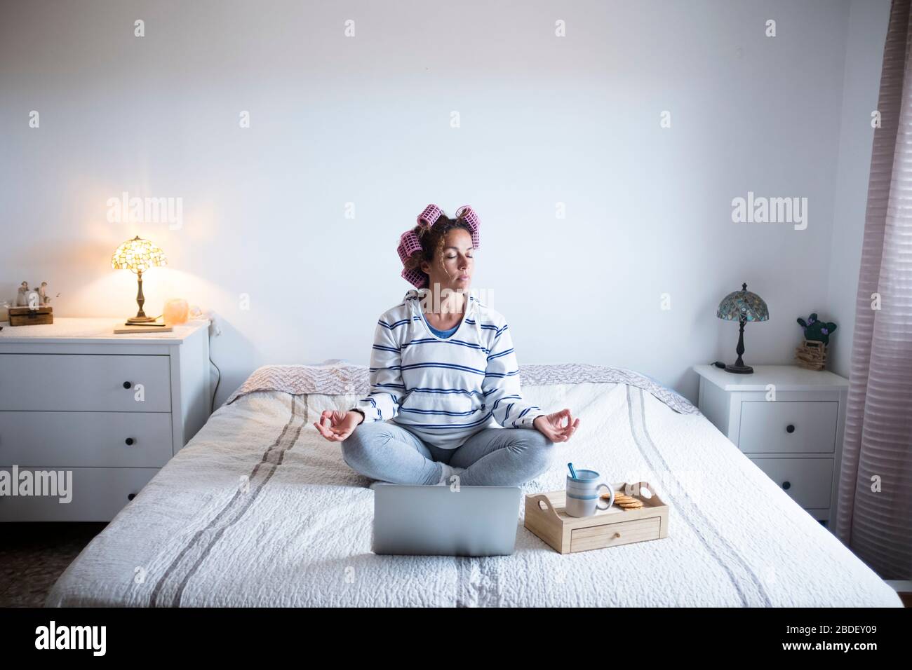 Woman with hair curlers meditating on bed in front of laptop Stock Photo