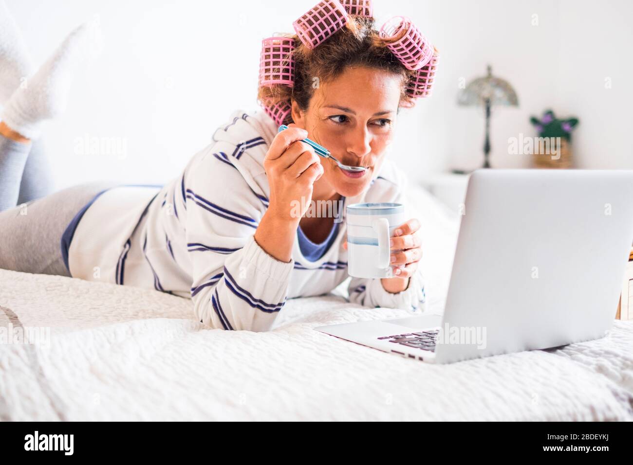 Woman with hair curlers lying on bed and looking at laptop Stock Photo