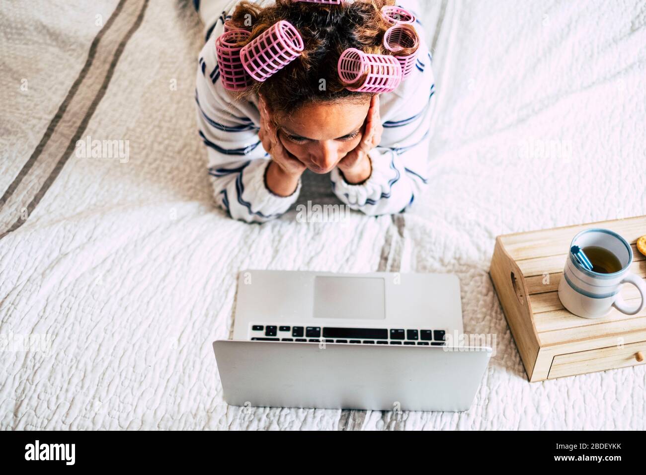 Woman with hair curlers lying on bed and looking at laptop Stock Photo