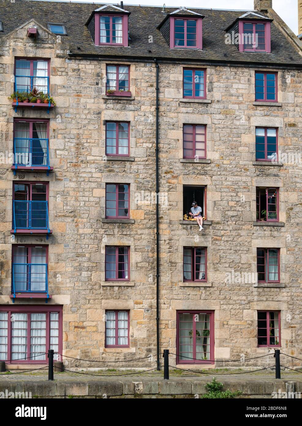 Leith, Edinburgh, Scotland, UK. 8th Apr, 2020. Covid-19 lockdown: on one of the warmest sunny days so far this year with people in lockdown. A man and his dog sit precariously in an open 3rd floor window in the Cooperage warehouse converted into flats on The Shore along the Water of Leith Stock Photo