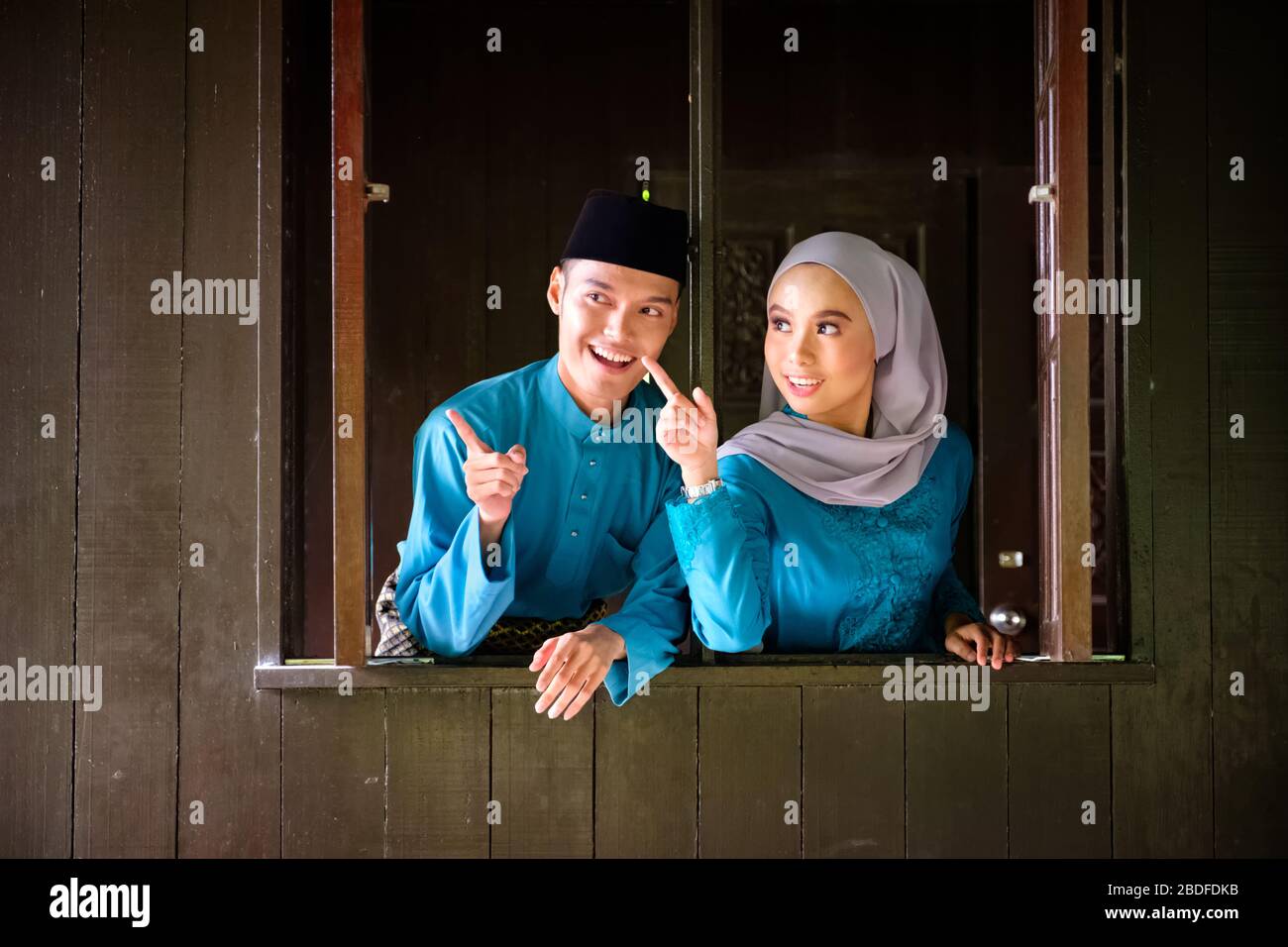 A portrait of young couple of malay muslim in traditional costume during Aidilfitri celebration at the wooden window of traditional house. Raya and Mu Stock Photo