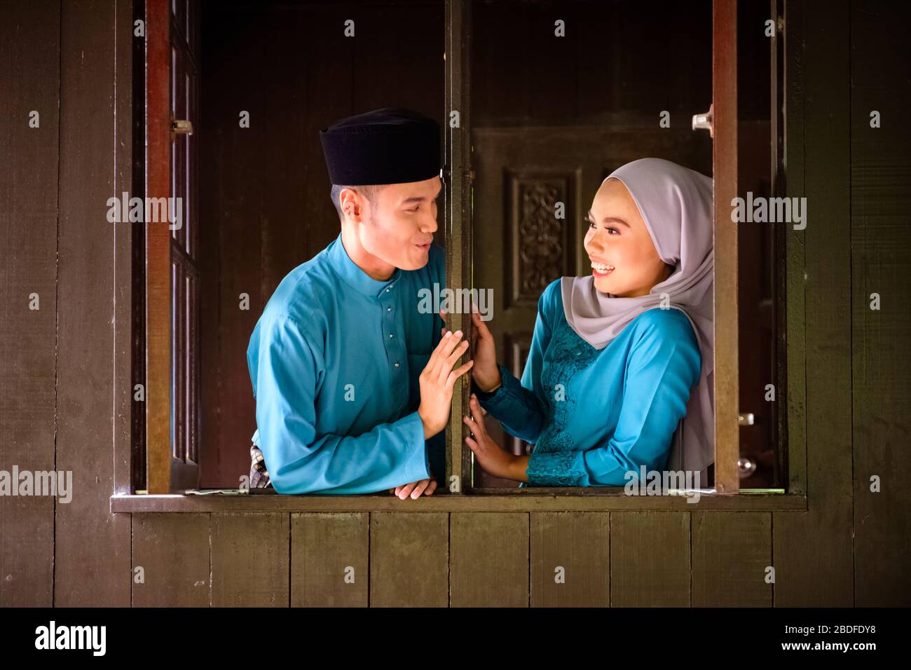 Young couple of malay muslim in traditional costume having sweet romantic conversation during Aidilfitri celebration at wooden window of traditional h Stock Photo