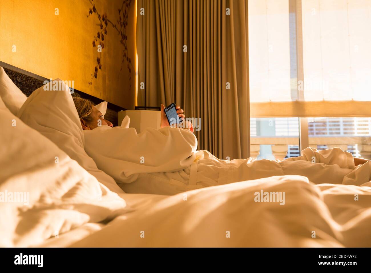 Adult woman executive lying on hotel bed in early morning sunlight, looking at smart phone Stock Photo