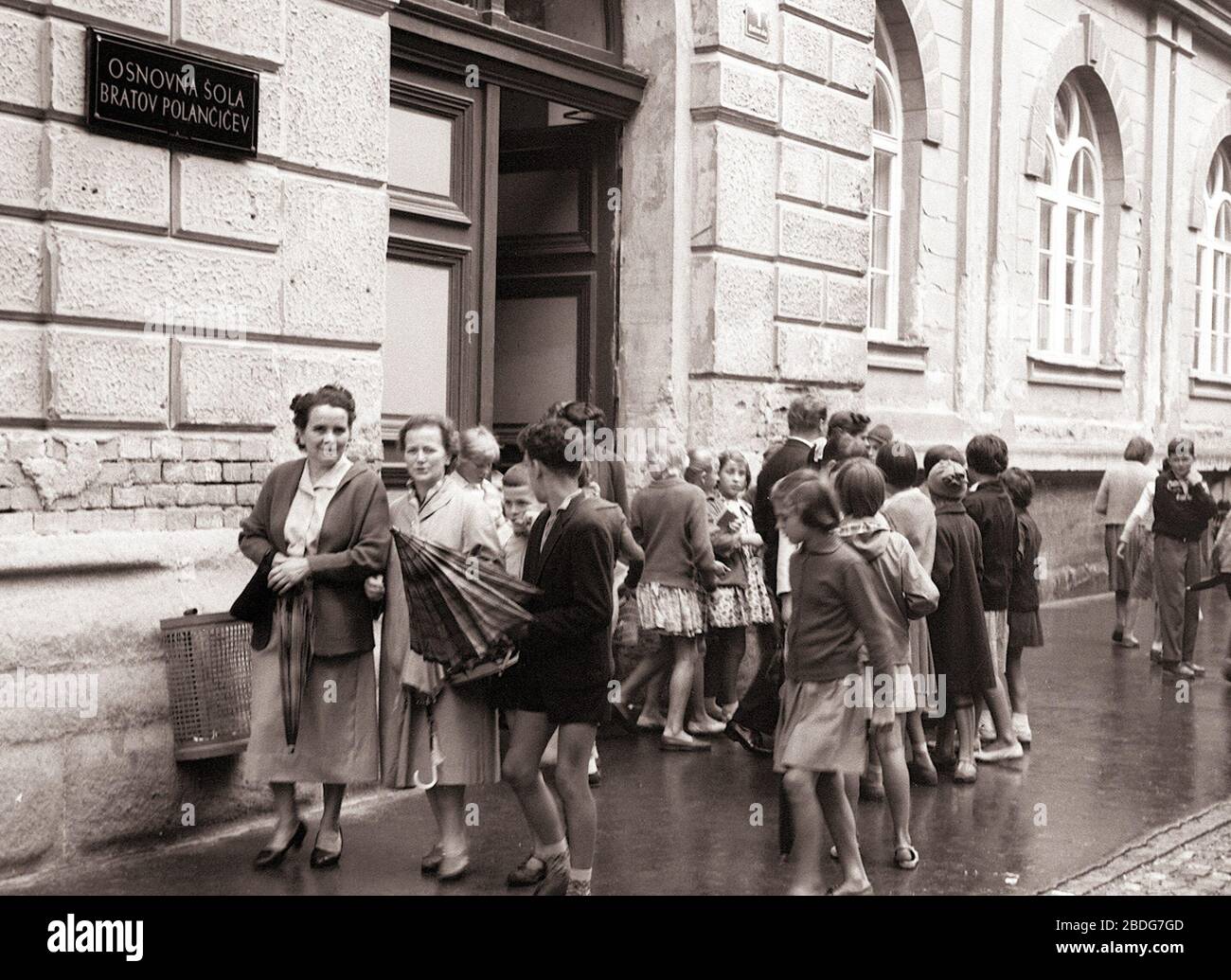 5. september 1960, Maribor: Po vseh ulicah in cestah našega mesta so se zlivali danes zjutraj skozi široko odprta šolska vrata potoki šolarjev. Najmanjše - tiste, ki gredo letos prvikrat v šolo - so na poti v šolo spremljale mamice, nekateri malčki pa so jo mahali prvikrat v šolo kar v spremstvu starejših bratov in sester. Tudi gost dež ni mogel šolarjem, ki so se v počitnicah do dobra naigrali, natekali in nakopali, pokvariti vznemirjenega veselja ob zopetnem srečanju s šolo. (avtor fotografije: Dragiša Modrinjak) Stock Photo