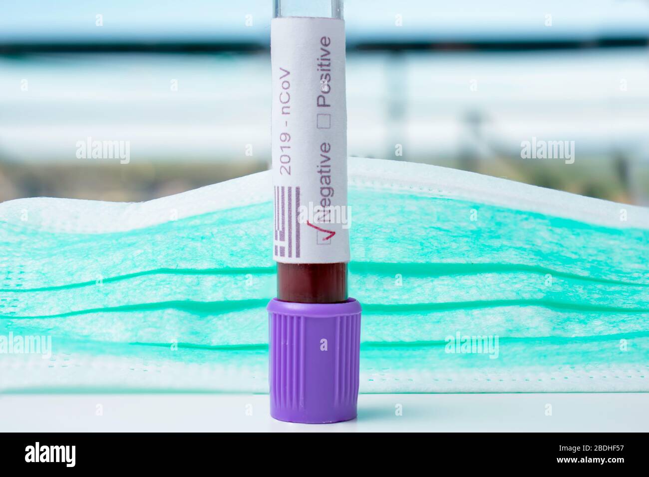 A  blood vial in front of a medical mask. The label on the vial states 2019 n Cov, the words positive negative. It also contains the Greek flag. Stock Photo
