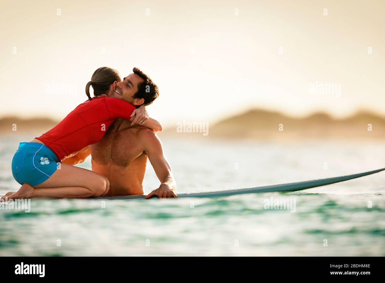 Woman kneeling on a surf-board, hugging her boyfriend. Stock Photo