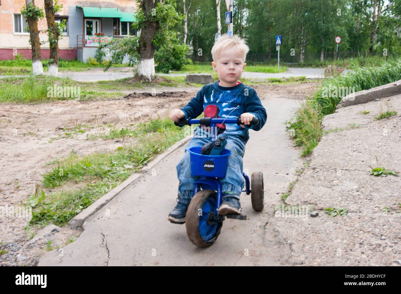Russia, Novodvinsk - July 2019. Blond-haired boy in blue rides a small tricycle on a broken city path Stock Photo