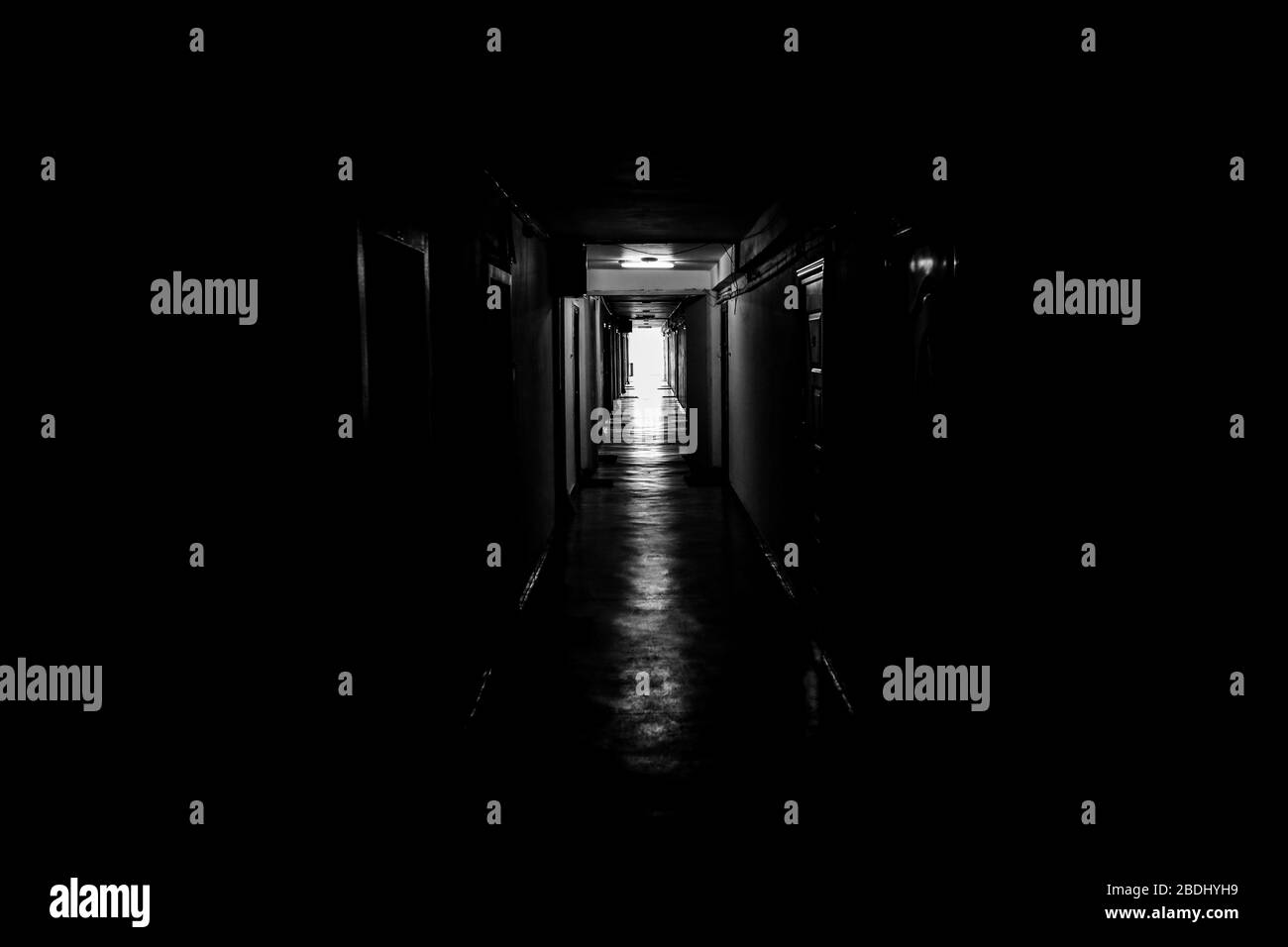 Black and white image with a dark mood hallway with apartment doors on each side - the light at the end of the tunnel. Stock Photo