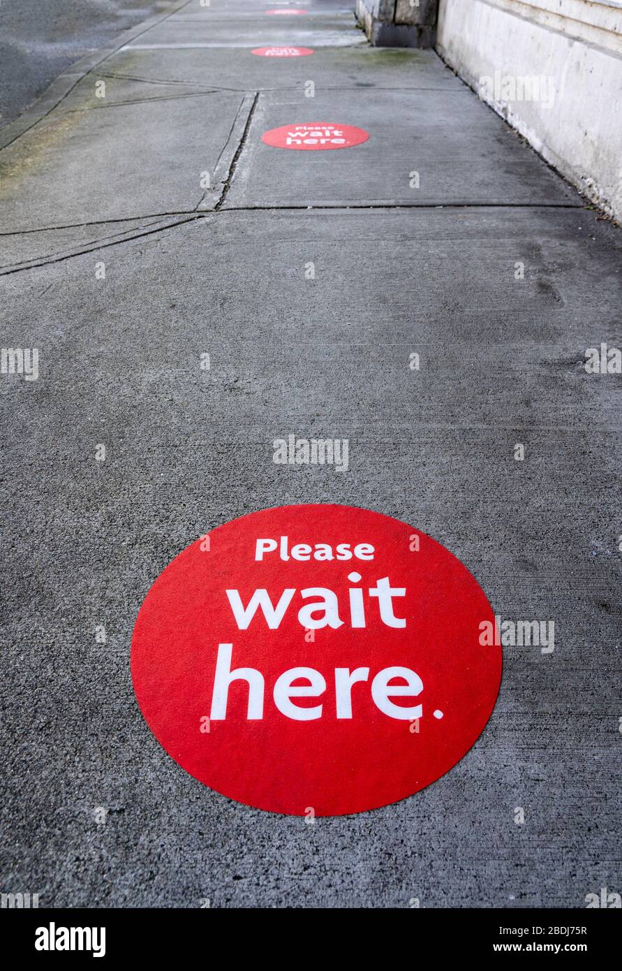 Round red physical distancing notification with words 'Please Wait Here' on sidewalk during Coronavirus Covid-19 outbreak Stock Photo