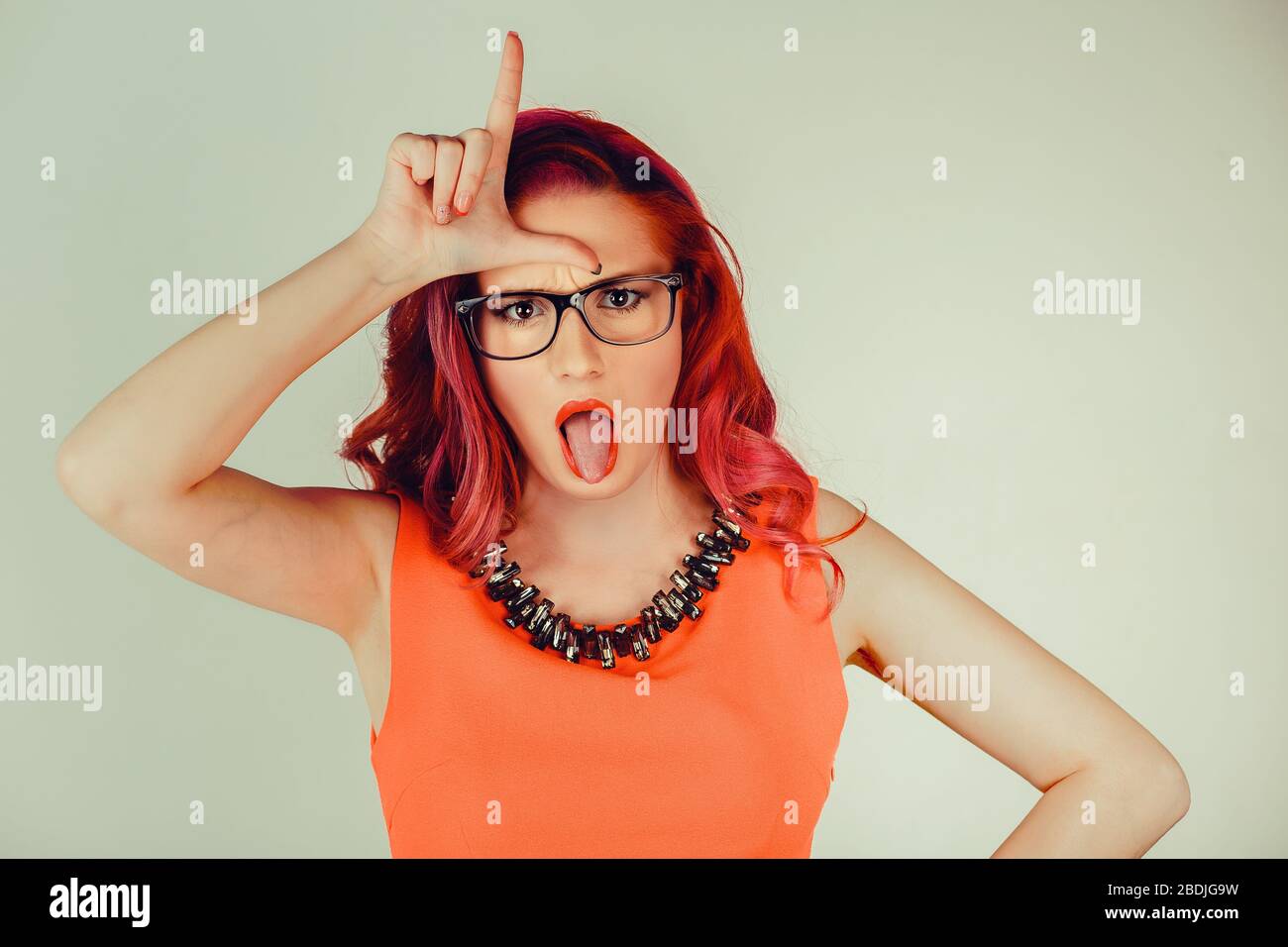 Closeup portrait young unhappy woman giving loser sign on forehead looking at you disgust on face isolated gray green wall background. Negative human Stock Photo