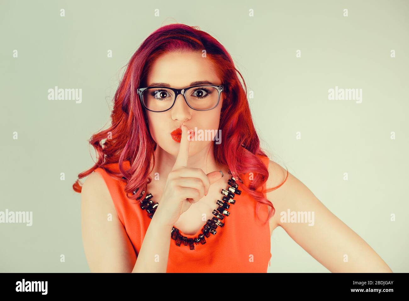 Woman wide eyed asking for silence or secrecy with finger on lips hush hand gesture green background wall. Pretty girl placing fingers on lips, shhh s Stock Photo