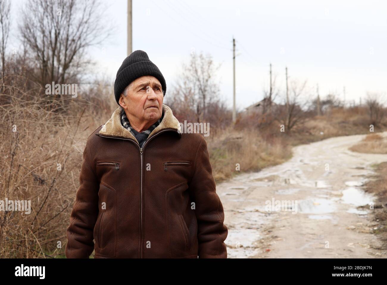 Elderly man standing on rural street in cloudy spring day. Worried face expression, concept of walking during coronavirus quarantine, life in village Stock Photo