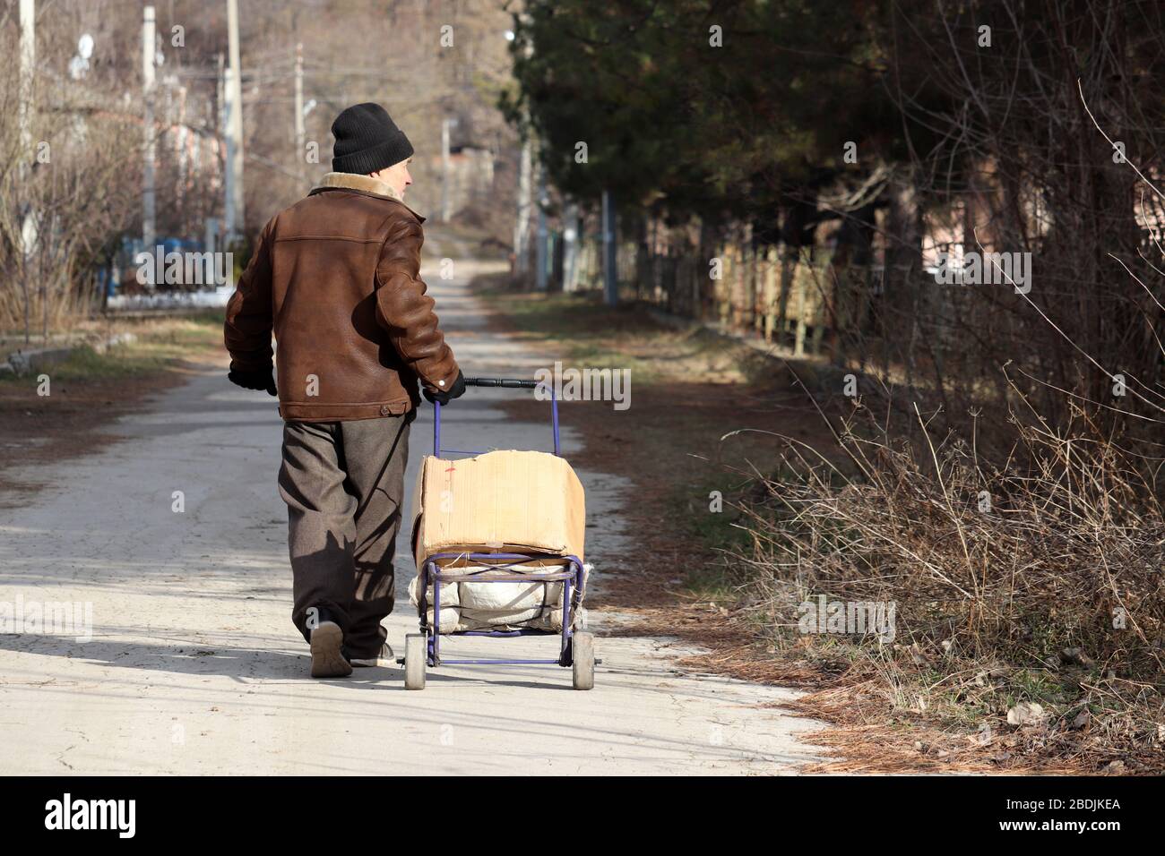 Elderly man walking on rural street with handcart, life in village. Retired person is stocking up on food during the quarantine of coronavirus Stock Photo