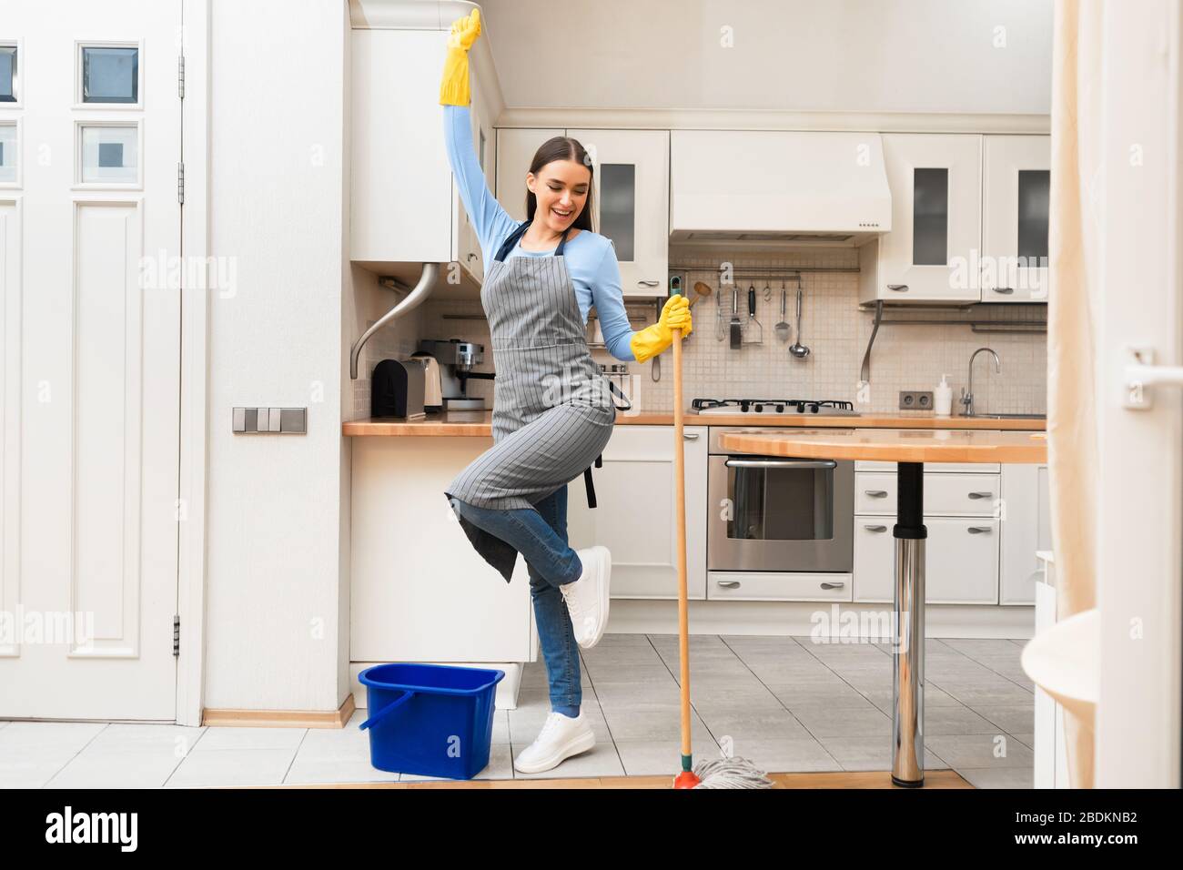 Happy young woman cleaning kitchen floor dancing with mop Stock Photo