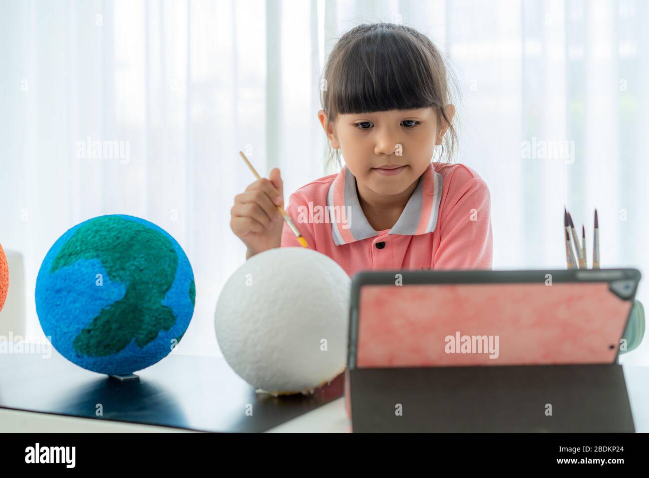 Asian elementary schoolgirl painting the moon in science class learning about the solar system via video conference with teacher and other classmates Stock Photo
